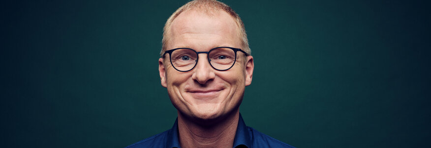 Hier sehen Sie ein Portraitfoto von dem #gesundkannjeder Experten Dr. Manfred Wagner. Er trägt ein blaues Hemd und ein Brille. Der Hintergrund ist einfarbig und dunkel. 