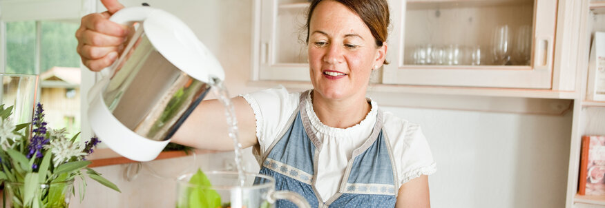 Hier sehen Sie eine Teekanne aus Glas in der viele frische Kräuter sind. Eine Frau schüttet heißes Wasser in die Teekanne und brüht den Tee auf. 