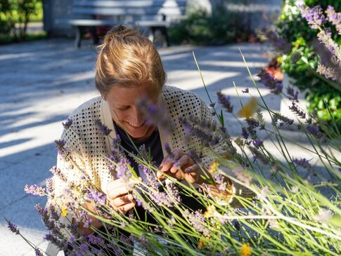 Hier sehen Sie eine Frau, welche in der Hocke ist und an Lavendelblüten riecht, welche am Wegesrand wachsen. 