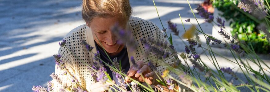 Hier sehen Sie eine Frau, welche in der Hocke ist und an Lavendelblüten riecht, welche am Wegesrand wachsen. 