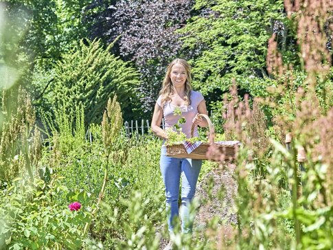 Hier sehen Sie eine junge Frau, die mit einem Korb voller Kräuter durch einen Kräutergarten spaziert.