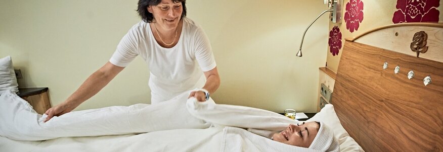 Hier sehen Sie eine Frau die von einer Therapeutin im Bett mit Decken und Tüchern komplett eingewickelt wird. Nur ihr Gesicht ist noch frei.