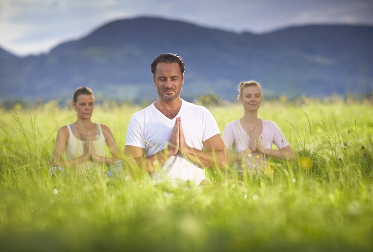 Hier sehen Sie eine Gruppe von drei Personen auf einer Wiese sitzen. Ein Mann sitzt vorne in der Mitte, hinter ihm sitzen links und rechts je eine Frau. Sie haben eine Meditationshaltung eingenommen, die Hände sind vor der Brust und die Augen sind geschlossen. Hinter ihnen erstreckt sich ein Bergpanorama. 