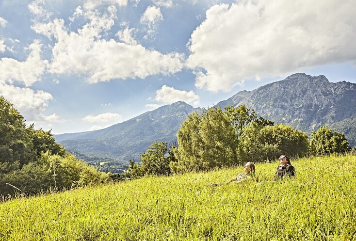 Hier sehen Sie zwei Frauen, die in einer dichten grünen Wiese vor einem Bergpanorama sitzen und die Sonne genießen.