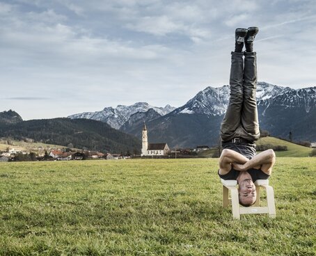Hier sehen Sie den Bayern Botschafter Kilian Trenkle. Auf einer Wiese macht er einen Kopfstand auf einem Yogahocker. Hinter ihm ist eine klassische bayerische Landschaft zu sehen.