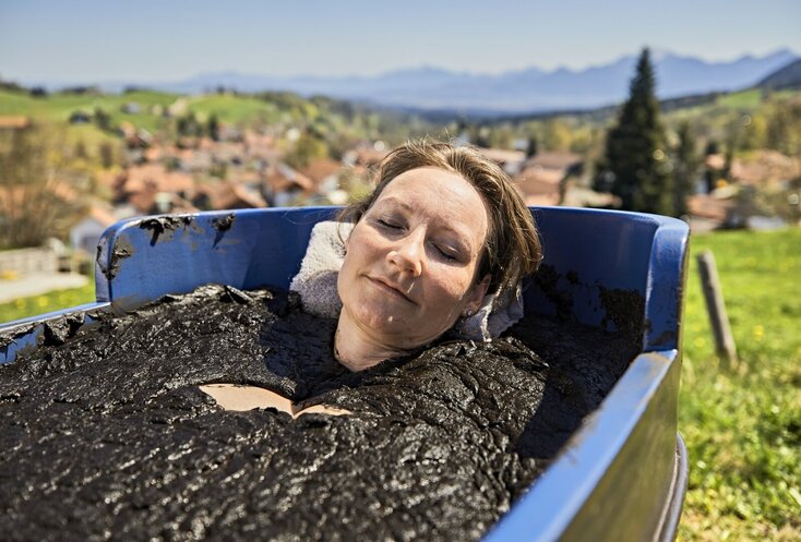 Sie sehen eine Frau, die in einer Holzwanne auf der Wiese ein Moorbad nimmt. Im Hintergrund ist der Ort Bad Kohlgrub zu sehen.