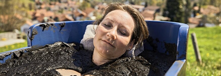 Sie sehen eine Frau, die in einer Holzwanne auf der Wiese ein Moorbad nimmt. Im Hintergrund ist der Ort Bad Kohlgrub zu sehen.