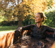 Frau sitzt mit Moor bedeckt bei einer Kur in einer hölzernen Badewanne draußen in der Herbstsonne