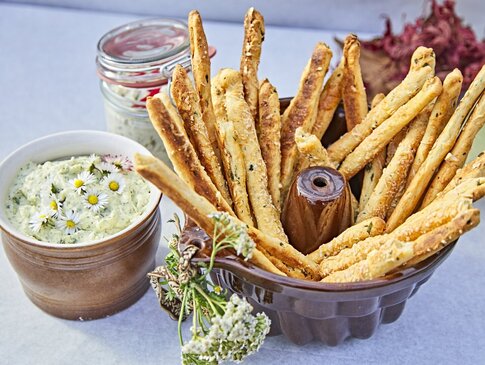 Hier sehen Sie auf einem Tisch eine kleine Schüssel mit selbstgemachtem frischen Kräuterquark. In einer größeren Schüssel daneben sind frische Teigstangen. Dekoriert ist der Tiasch mit Kräutern und Blüten.