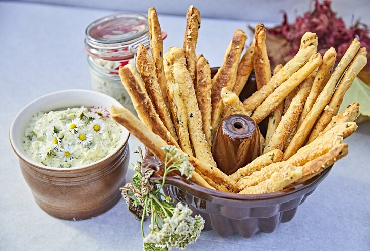 Hier sehen Sie auf einem Tisch eine kleine Schüssel mit selbstgemachtem frischen Kräuterquark. In einer größeren Schüssel daneben sind frische Teigstangen. Dekoriert ist der Tiasch mit Kräutern und Blüten.