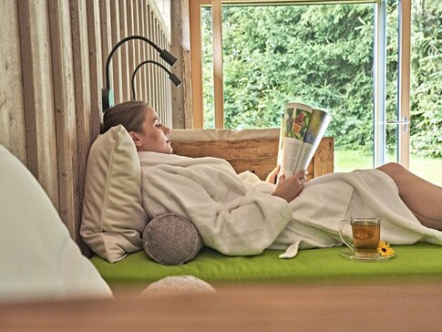 Hier sehen Sie eine Frau in einem weißen Bademantel beim Lesen auf einer Liege nahe bei einem großen Fenster liegen, mit Blick auf einen grünen Garten.