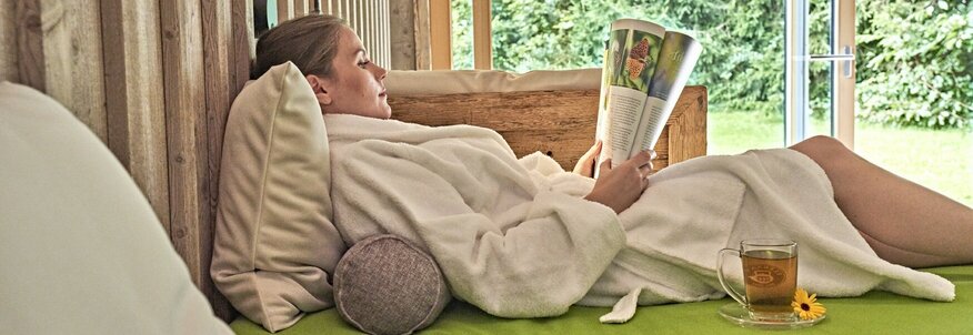 Hier sehen Sie eine Frau in einem weißen Bademantel beim Lesen auf einer Liege nahe bei einem großen Fenster liegen, mit Blick auf einen grünen Garten.