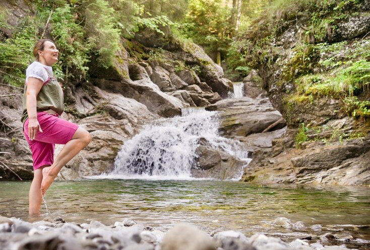 Hier sehen Sie einen kleinen Bergfluss. Bayern Botschafterin Christine Waibel-Beer steht an einer seichten Stelle im Wasser und kneippt im Storchenschritt. 