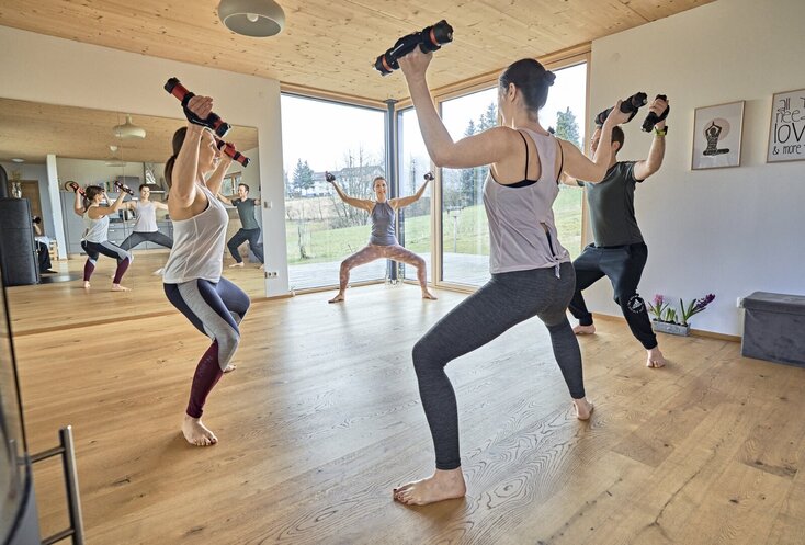 Drei Frauen und ein Mann machen Yoga-Übungen mit Gewichten in den Händen. Sie stehen im Kreis auf einem Holzfußboden, haben die Beine breit aufgestellt und strecken die Hände mit den Hanteln angewinkelt in die Luft. Ein großer Spiegel ist an der Wand gegenüber und durch die großen Fenster sieht man nach draußen auf die Wiese und zu den Bäumen.