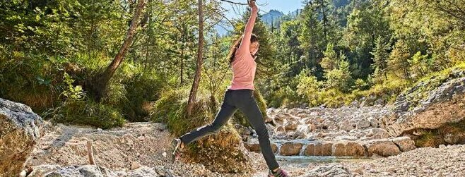 Hier sehen Sie einen kleinen Flusslauf in Mitten von Bäumen und Büschen. Eine Frau springt über diesen Flusslauf von einem Stein zum anderen Stein.