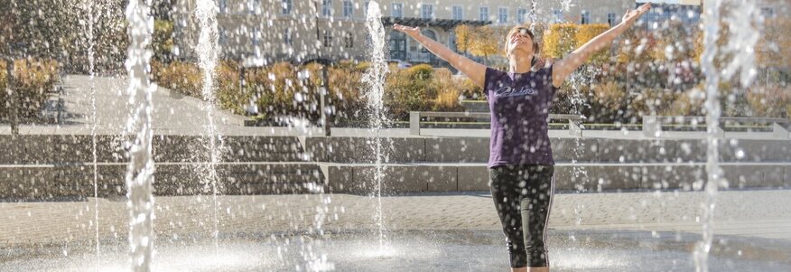 Hier sehen Sie eine Frau in einem Kurpark. Sie steht in mitten eines Springbrunnen. Das Wasser steigt von unten nach oben und die Frau lacht und hebt ihre Hände. 