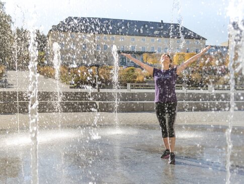 Hier sehen Sie eine Frau in einem Kurpark. Sie steht in mitten eines Springbrunnen. Das Wasser steigt von unten nach oben und die Frau lacht und hebt ihre Hände. 