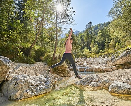 Hier sehen Sie einen kleinen Flusslauf in Mitten von Bäumen und Büschen. Eine Frau springt über diesen Flusslauf von einem Stein zum anderen Stein.