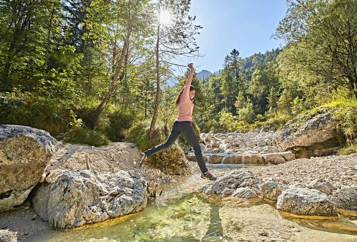 Hier sehen Sie einen kleinen Flusslauf in Mitten von Bäumen und Büschen. Eine Frau springt über diesen Flusslauf von einem Stein zum anderen Stein.