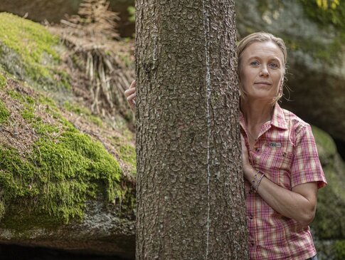 Hier sehen Sie die Bayern Botschafterin und Waldbademeisterin Elke Seidel. Sie steht seitlich an einem Baum.