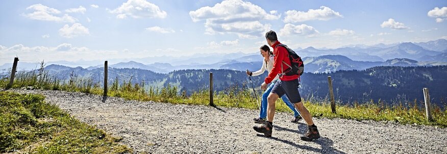 Hier sehen Sie zwei Wanderer, die auf einem Wanderweg in den Bergen gehen und das Panorama über grüne Wiesen und Wälder im Tal genießen.