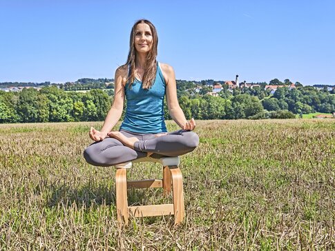 Hier sehen Sie eine junge Frau, die im Schneidersitz auf einem Holzhocker in einem Feld sitzt und meditiert.