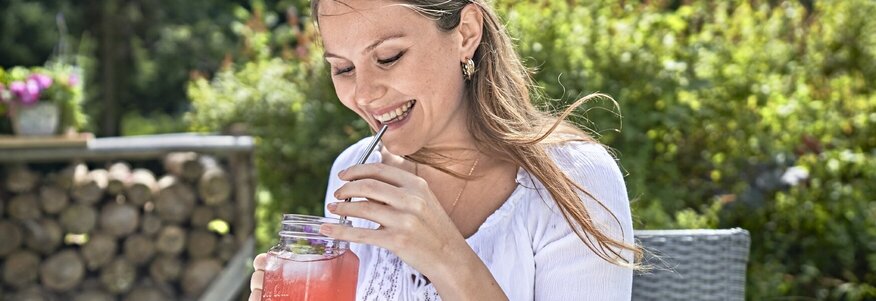 Hier sehen Sie eine junge Frau, die lächelnd draußen im Garten sitzt und Fruchtsaft trinkt. 