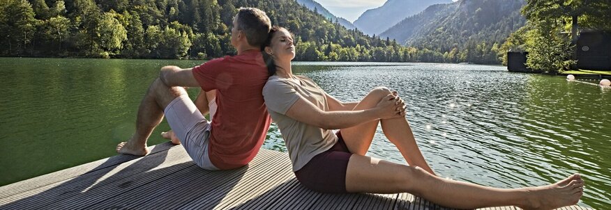 Sie sehen einen Mann und eine Frau mit dem Rücken aneinandergelehnt auf einem Steg am See. Im Hintergrund sehen Sie Berge.