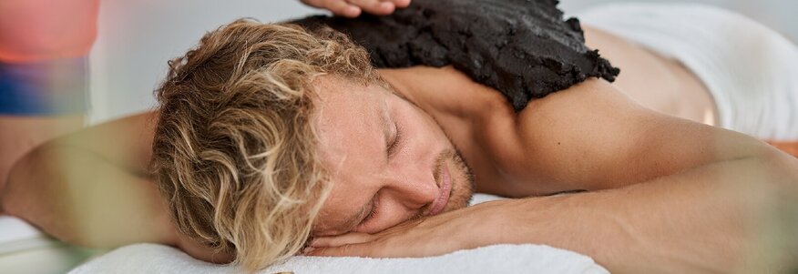 Hier sehen Sie einen Mann, der mit dem Bauch auf einer Massageliege liegt. Er bekommt von einer anderen Person eine Moorpackung auf den Rücken gestreicht. 