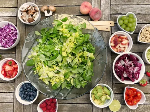 Hier sehen Sie eine große Schüssel mit grünen Salat. Um die Schüssel stehen viele kleine Schüsseln mit Zutaten, die man dem Salat hinzu geben kann. Zum Beispiel Tomaten, Nüsse, Zwiebeln, Beeren und noch vieles mehr. 