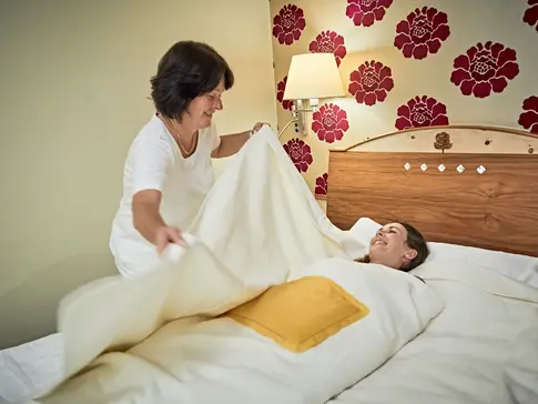 Hier sehen Sie eine Frau, welche eine Schrothkur Behandlung bekommt. Sie liegt in einem Bett und wird von einer Therapeutin mit mehreren Decken und Wärmekissen zugedeckt. 
