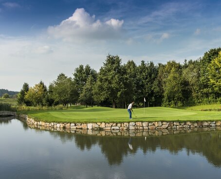 Auf dem Bild sehen Sie einen Golfer auf saftig grünem Rasen an einem Fluss. Im Hintergrund sind Bäume und der strahlend blaue Himmel zu sehen.