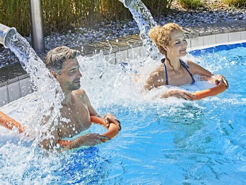 Hier sehen Sie einen Mann und eine Frau in einem Außenpool. Beide Personen stehen unter einem Wasserfall, welcher auf den Schultern der Personen landet. 