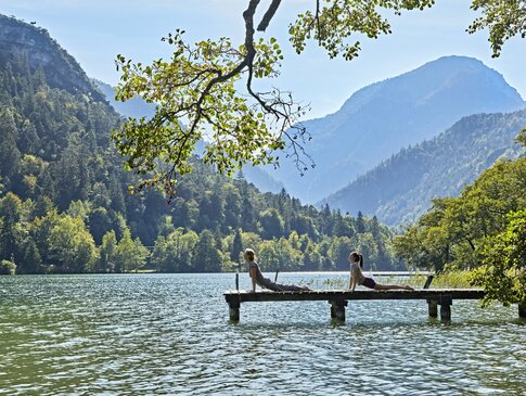 Hier sehen Sie Personen, die auf einem Steg Yoga praktizieren. Hinter ihnen sind die Berge zu sehen. 