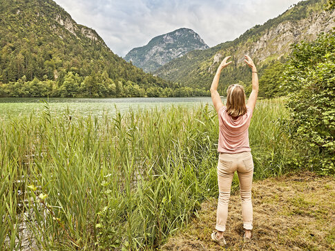 Hier sehen Sie eine Frau von hinten. Sie hat ihre Arme über die Seite nach oben über den Kopf gehoben. Sie blickt auf eine See- und Berg Landschaft.