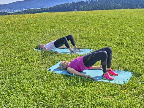 Hier sehen Sie zwei Frauen auf einer Wiese. Sie tragen Sportkleidung und machen Rückenübungen auf den Boden.