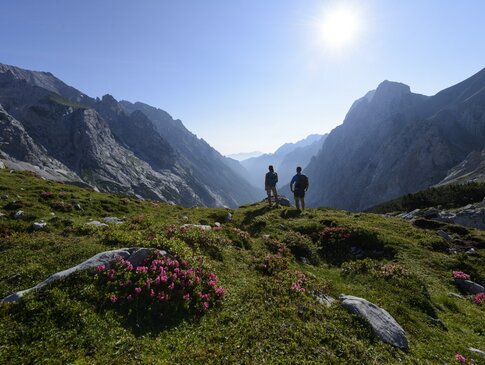 Hier sehen Sie zwei Personen beim Wandern auf den Bergen. Sie stehen mit dem Rücken zu uns und Blicken auf die Berge. 