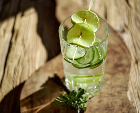 Hier sehen Sie ein Glas Wasser, welches auf einem Holzbrett steht und sowohl Gurken als auch Limetten-Scheiben beinhaltet.