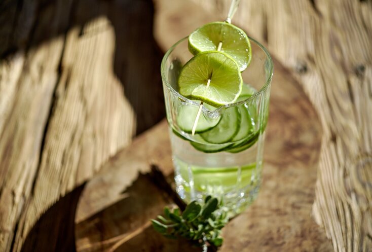 Hier sehen Sie ein Glas Wasser, welches auf einem Holzbrett steht und sowohl Gurken als auch Limetten-Scheiben beinhaltet.