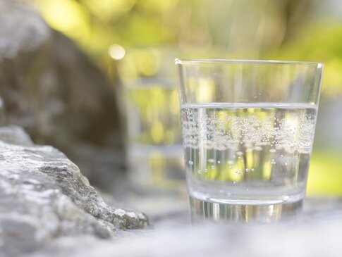 Hier sehen Sie ein Glas mit Heilwasser gefüllt. Das Glas steht im Freien auf einem Stein. Der Fokus liegt auf dem Glas, der Rest ist unschwarf. 