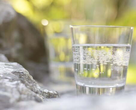 Hier sehen Sie ein Glas mit Heilwasser gefüllt. Das Glas steht im Freien auf einem Stein. Der Fokus liegt auf dem Glas, der Rest ist unschwarf. 