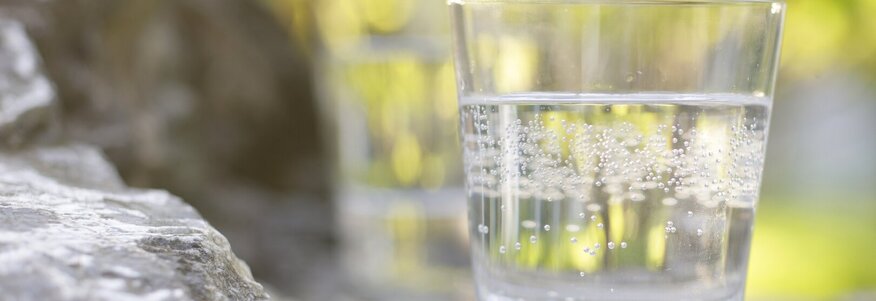 Hier sehen Sie ein Glas mit Heilwasser gefüllt. Das Glas steht im Freien auf einem Stein. Der Fokus liegt auf dem Glas, der Rest ist unschwarf. 