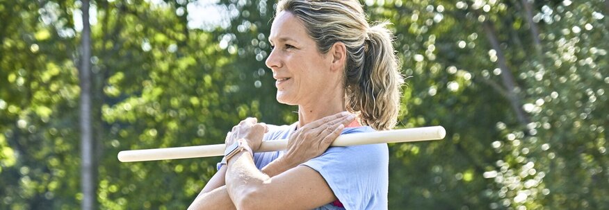 Ich sehe eine Frau mit schwarzer Sporthose und hellblauem T-Shirt auf einer Bank sitzen. Mit Hilfe eines Holzstabes dehnt Sie durch Drehung des Oberkörpers ihren Rumpf. 