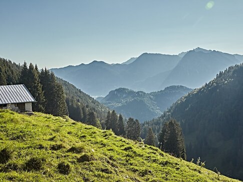 Hier sehen Sie eine Berglandschaft mit einer grünen Wiese davor. Eine kleine Alm ist auf der linken Seite zu sehen.