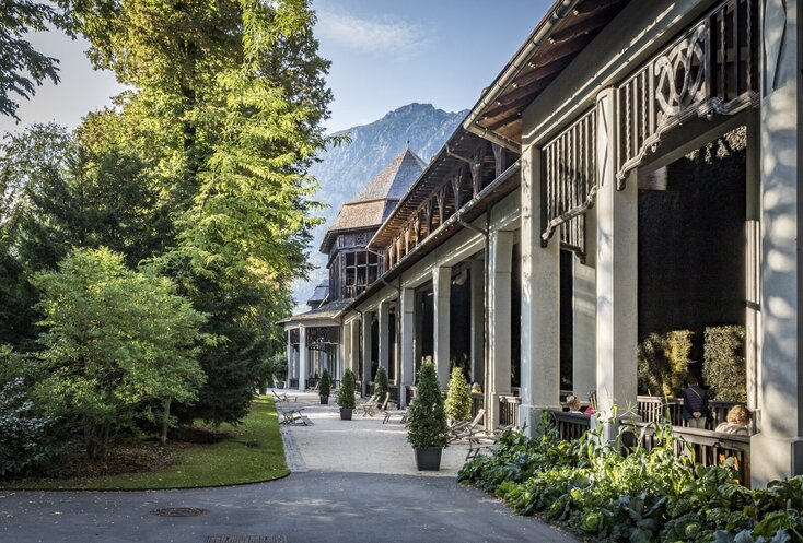 Rechts im Bild ist das AlpenSole Gradierhaus in Bad Reichenhall zu sehen. Dahinter ein mächtiger Berggipfel und auf der linken Seite Bäume. Im Gradierhaus in Bad Reichenhall, kann man die AlpenSole inhalieren.