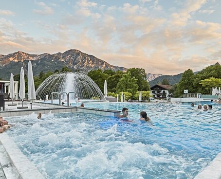 Hier sehen Sie eine Pool im Außenbereich. Es sind vereinzelnt Menschen in dem Pool und liegen auf den Massagedüsen. Im Hintergrund sind Berge zu sehen. 