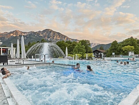 Hier sehen Sie eine Pool im Außenbereich. Es sind vereinzelnt Menschen in dem Pool und liegen auf den Massagedüsen. Im Hintergrund sind Berge zu sehen. 