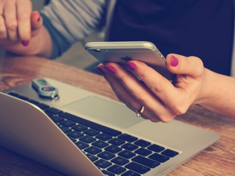 Eine Dame sitzt vor Ihrem Laptop mit dem Mobiltelefon in der linken Hand.