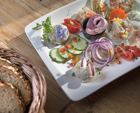 Hier sehen Sie einen Brotzeitteller mit viel Gemüse, wie Gurke und Tomate, ebenso sehen Sie unterschiedliche Brotaufstriche und dunkles Bauernbrot. 