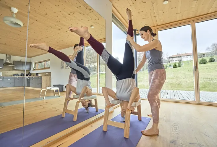 GESUNDES BAYERN-Expertin Susanne Lindlbauer hilft einer Dame bei einer Yoga-Übung auf einem Kopfstandhocker und hält dabei ihre Beine. Der Yoga-Stuhl steht auf einer lilafarbenen Matte neben einem großen Spiegel. Sie trägt eine enge rosa Hose und ein lilafarbenes Top, die Dame auf dem Stuhl trägt eine enge schwarze Hose mit grauen und roten Streifen und ein graues T-Shirt. Durch das große Fenster hinter den beiden Damen sind eine grüne Wiese mit Bäumen und der blaue Himmel zu sehen.